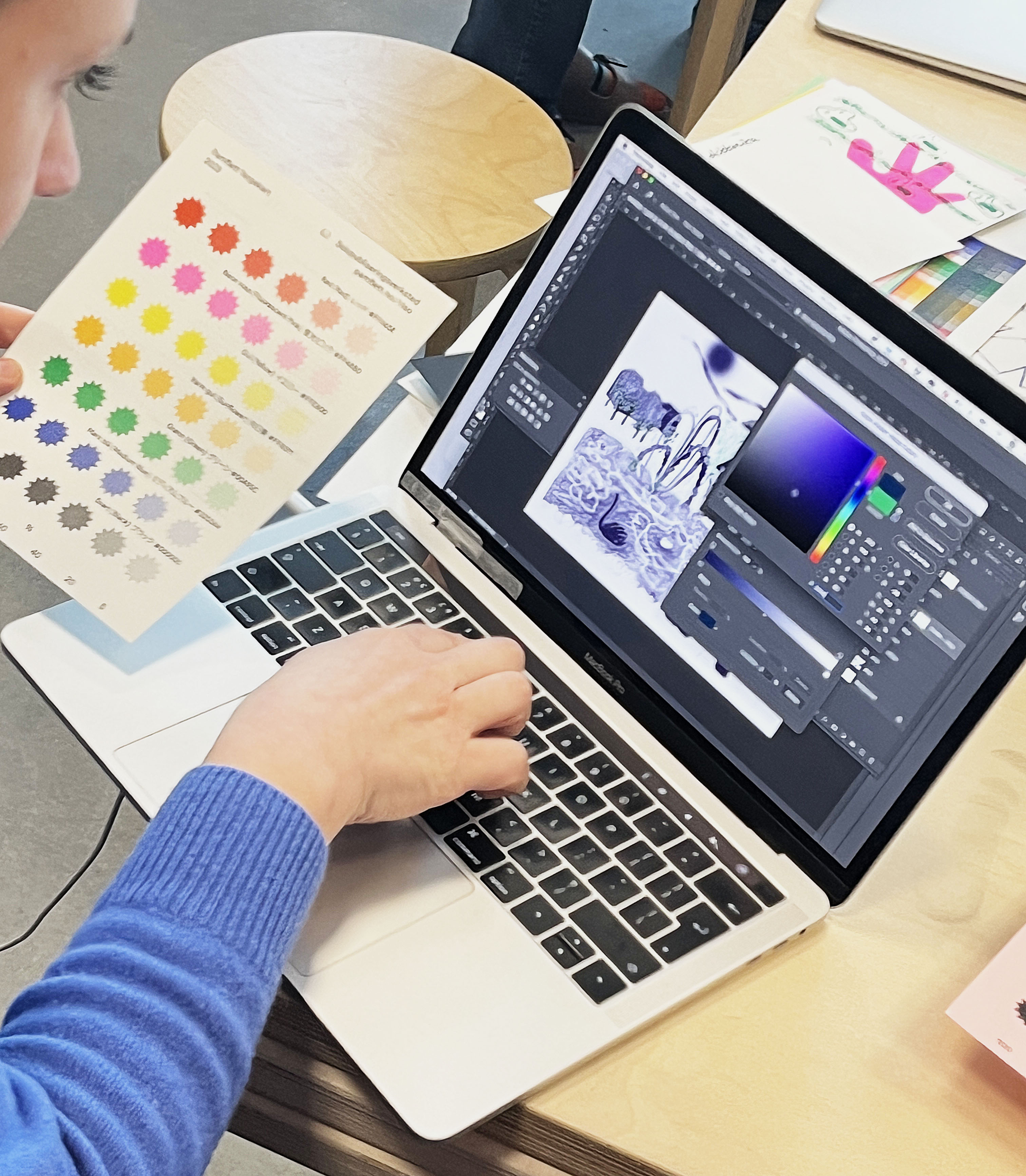 A white person in a blue sweater is holding a color swatch print, while typing on a computer keyboard with their other hand. On the computer screen Photoshop is open.