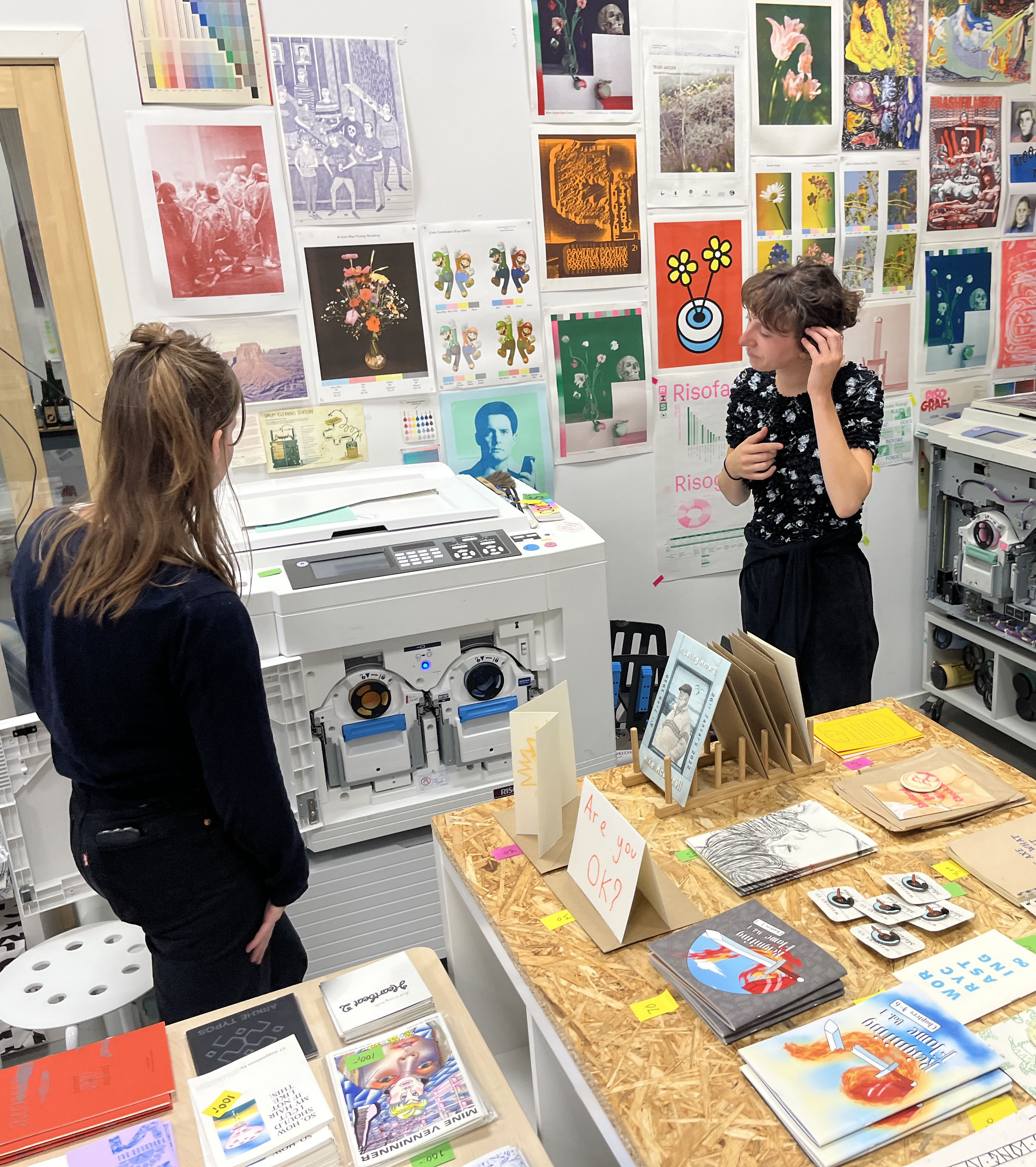 Two people are standing in our workshop next to a risograph printer, looking at the printer. In the background is our wall, filled with risoprinted posters. In the foreground there are two tables with several riso printed zines and prints.